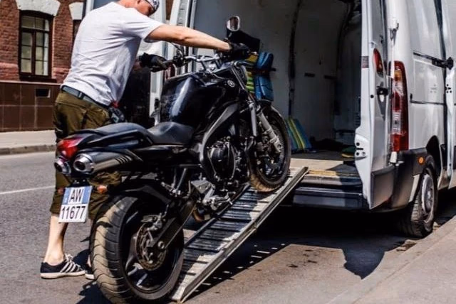 A man loads a black motorcycle onto a white cargo van using a metal loading ramp on a city street.